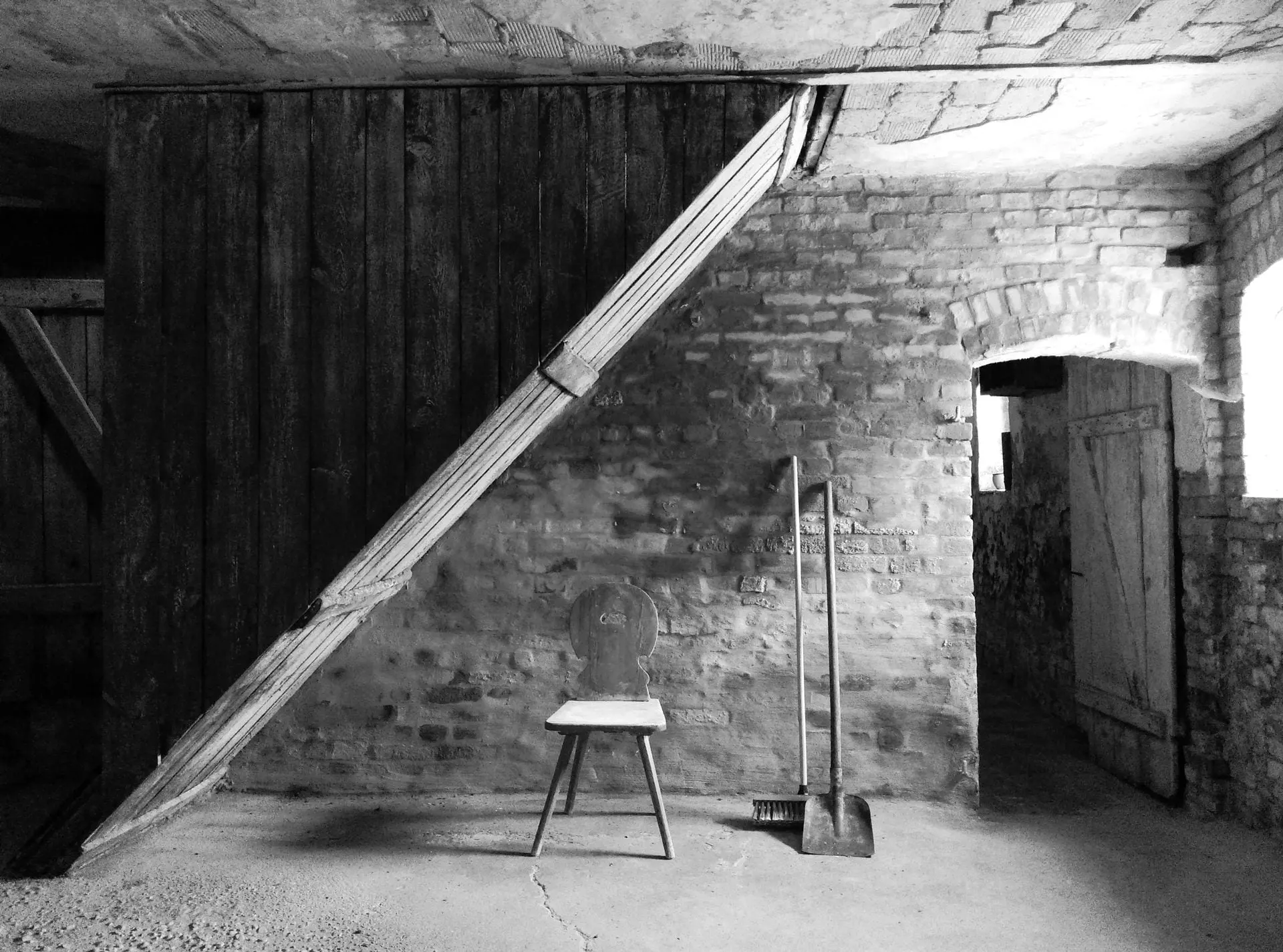 Schwarz-weisses Interieur mit Holztür, schrägem Stützbalken, Hocker, Schaufel und Ziegelwänden.