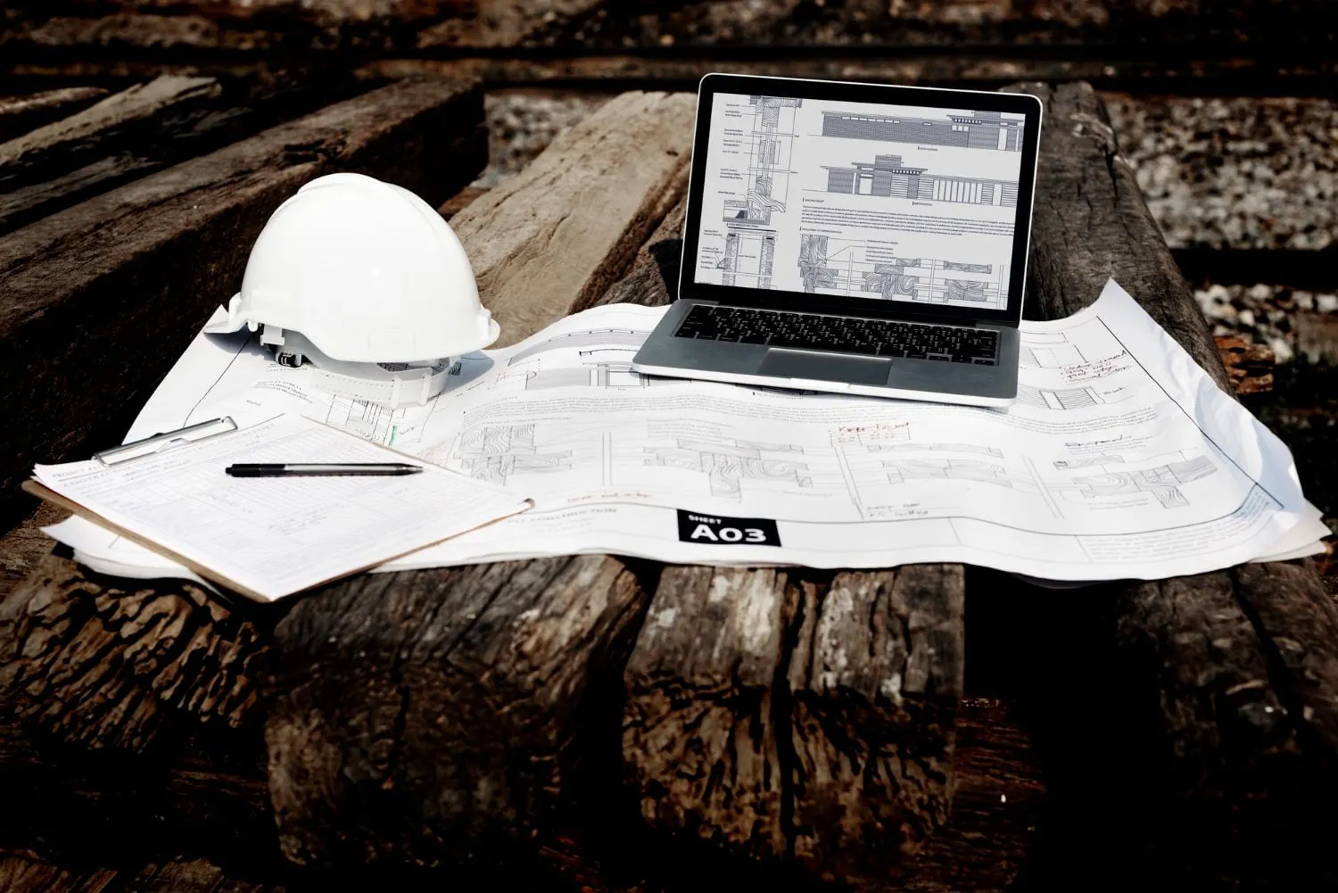 Weisser Schutzhelm, Laptop und Baupläne auf Holzbalken.