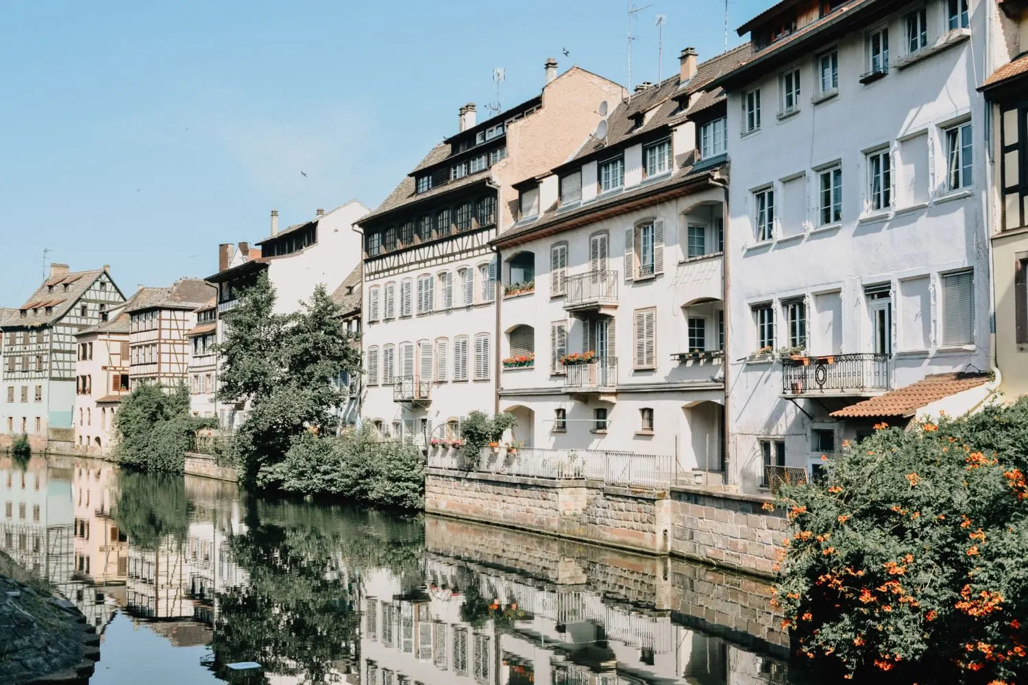 Gebäude entlang eines Kanals in Strassburg, Frankreich; helle Fassaden, Spiegelungen im Wasser, blauer Himmel.