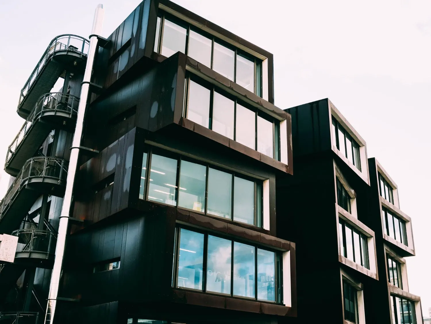 A low-angle view of a modern, multi-story building with dark, box-like modular sections and large windows.